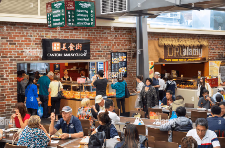 Busy indoor food court with people ordering at Canton-Malay Cuisine and Tropical Indian and Sri Lankan Cuisine stalls, with others seated at tables eating.