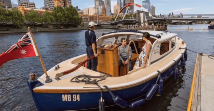 Small blue and white boat with three people onboard floating on a river near a city bridge.