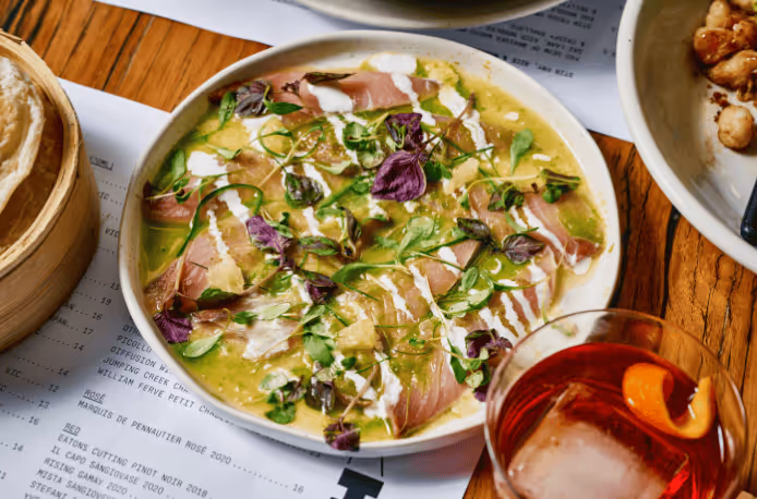 Plate of sashimi-style fish garnished with herbs and drizzled with white sauce, next to a glass with an orange peel and ice.