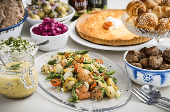 Table set with various dishes including a vegetable salad with asparagus and cherry tomatoes, meatballs, baked pie, red cabbage salad, and baked bread rolls.
