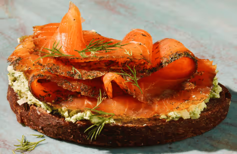 Open-faced sandwich with dark rye bread topped with herb cream cheese, smoked salmon slices, and dill sprigs.