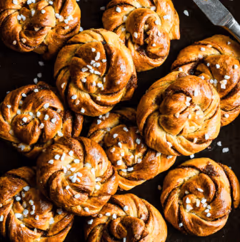 Close-up of cinnamon buns topped with pearl sugar on a dark surface.