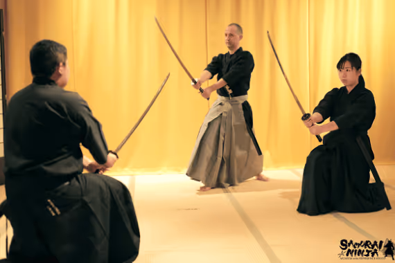 Three martial artists practicing with katanas in traditional uniforms on a light floor with a yellow curtain background.