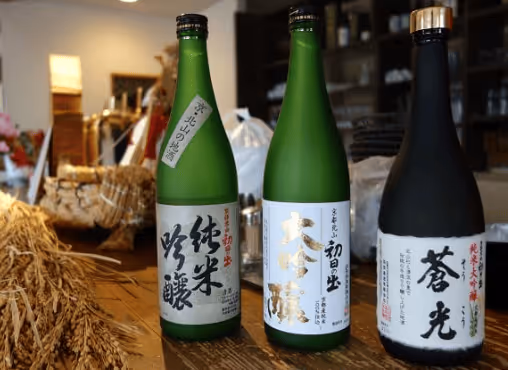 Three bottles of Japanese sake with labels in Japanese characters placed on a wooden surface, with dried rice plants beside them.