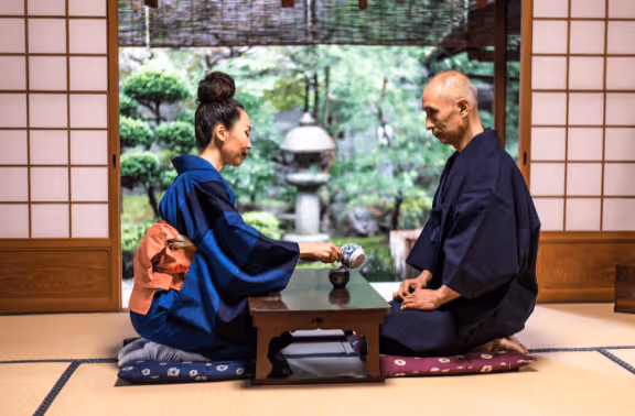 A woman in a blue kimono serving tea to a man in a dark kimono during a traditional Japanese tea ceremony in a room with tatami mats and shoji screens.