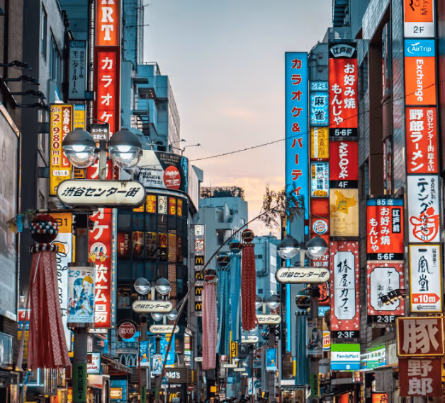 Bustling urban street in Japan at dusk with numerous colorful vertical neon signs and lanterns lining tall buildings.