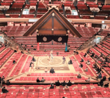 Sumo wrestling match taking place in a traditional indoor arena with sparse spectators seated in red chairs around the ring.
