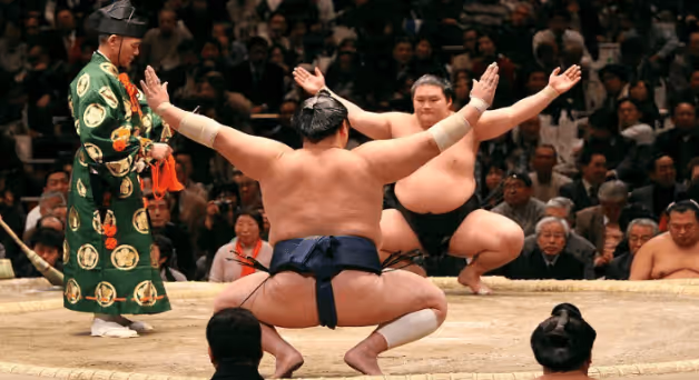 Two sumo wrestlers squatting and facing each other in a traditional ring with a referee standing nearby and an audience watching.
