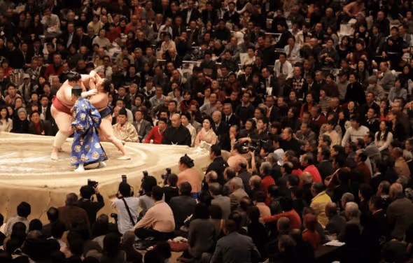 Two sumo wrestlers grappling inside a ring with a referee in a blue traditional outfit and a large audience watching.