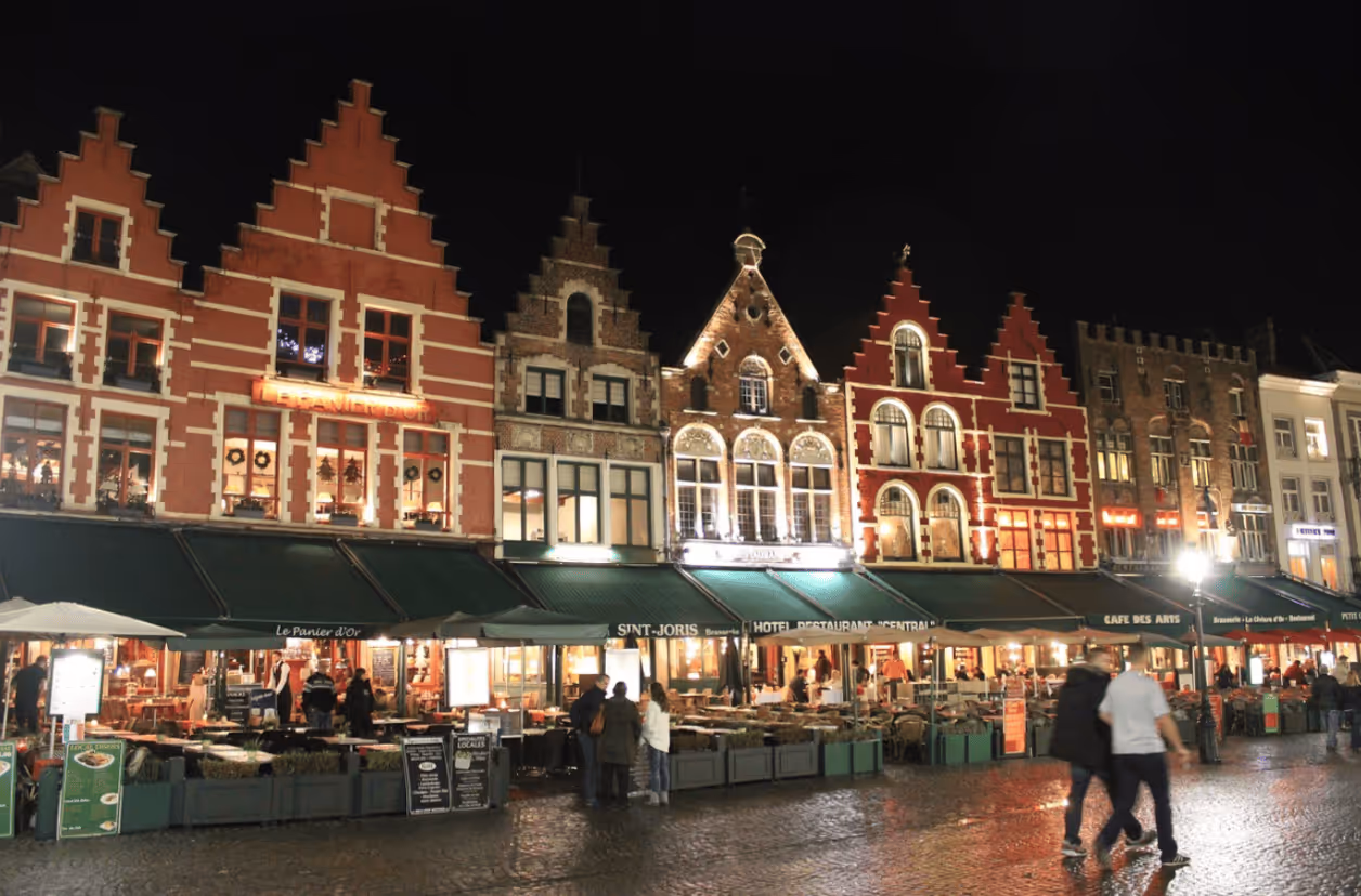 Night view of a row of historic stepped-gable buildings with lit restaurants and cafes in front, people walking on cobbled street.