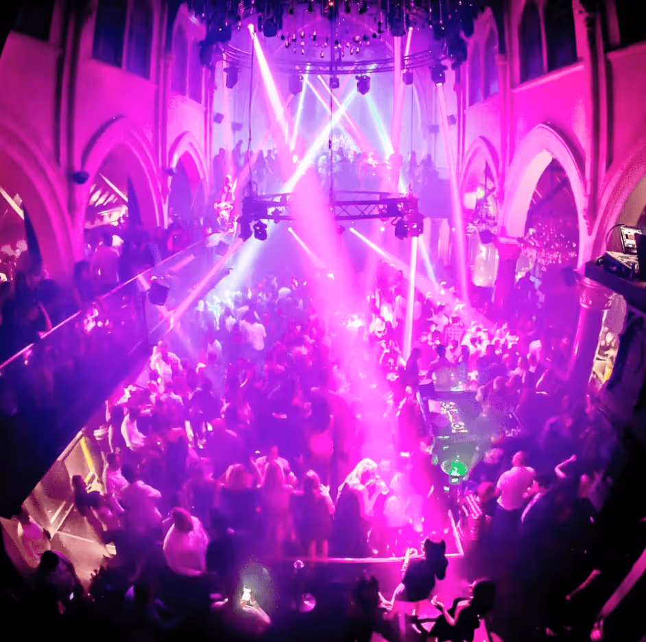 Crowded nightclub with purple and pink lighting and beams, featuring people dancing and a DJ setup.