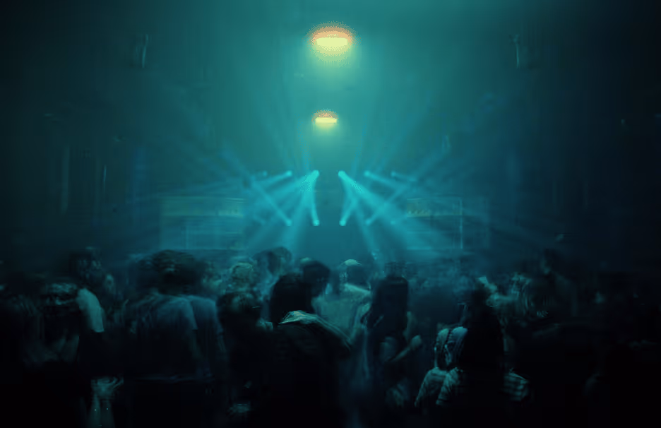 Crowd of people in a dimly lit indoor venue with teal stage lights shining above.