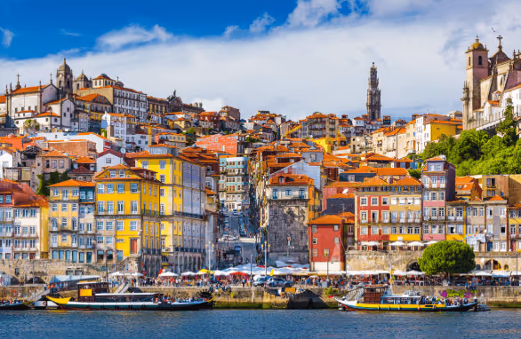 Colorful historic buildings with red rooftops along a waterfront, boats on the water, and a blue sky with clouds.
