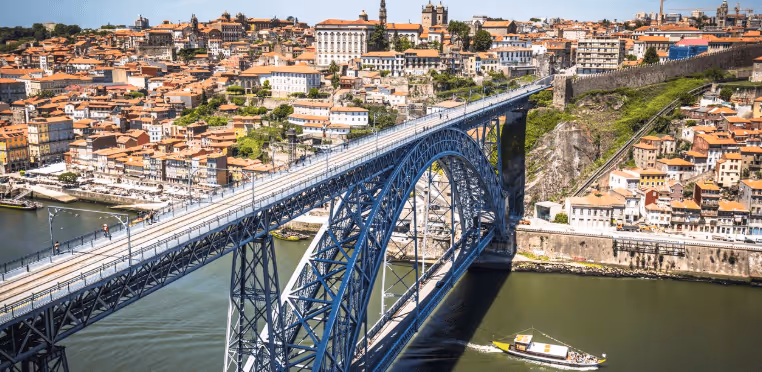 Blue arched metal bridge over a river with a boat passing underneath and a city with terracotta-roofed buildings in the background.