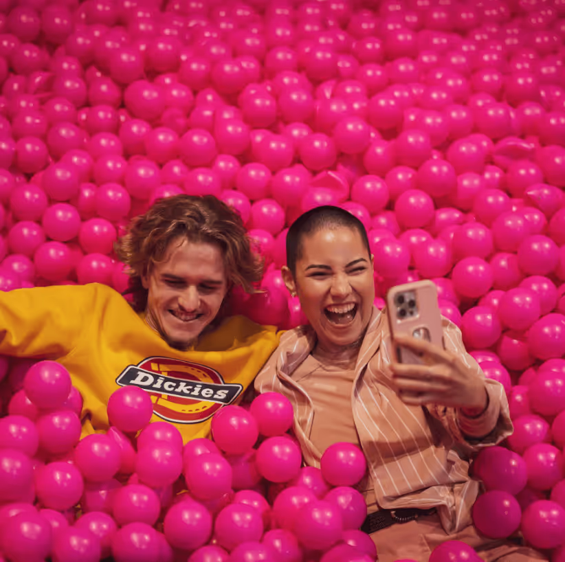 Two people smiling and taking a selfie while lying in a ball pit filled with bright pink balls.