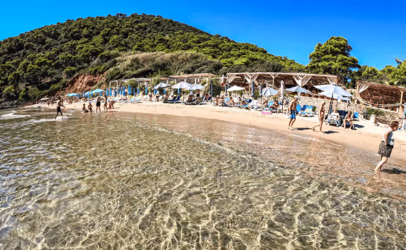 Sunny beach with clear shallow water, people walking and lounging under umbrellas near green wooded hill.