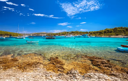 Clear turquoise water with rocky shore in foreground, boats anchored near green tree-lined coast under blue sky with scattered clouds.