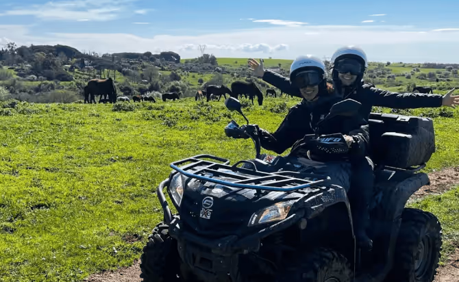 Two people wearing helmets riding a black ATV on a dirt path with green fields and grazing horses in the background.