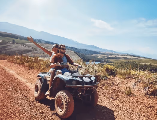 Man and woman riding a quad bike on a dirt trail through a scenic mountainous landscape under a partly cloudy sky.