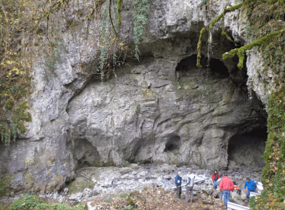 Group of people exploring the entrance of a large rocky cave surrounded by green moss and vegetation.