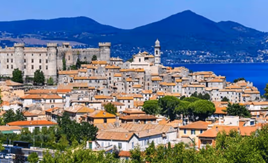 Scenic view of a coastal town with terracotta-roofed buildings, a historic castle, and mountains in the background under a clear blue sky.