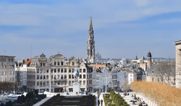 View of a European cityscape with historic buildings, a tall cathedral spire, and a statue in a formal garden with trimmed hedges and people walking.