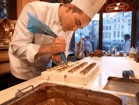 Chef in white uniform and hat carefully decorating chocolates with a piping bag in a kitchen.
