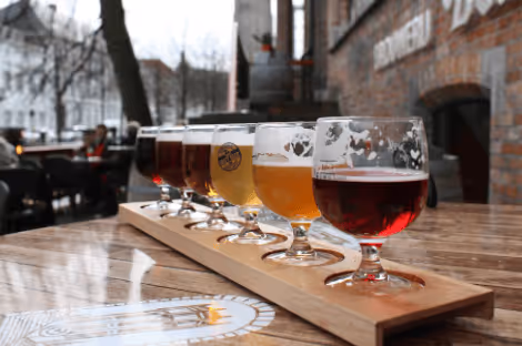 Flight of assorted craft beers in small glasses on a wooden paddle on a pub table outdoors.