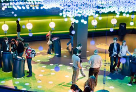 People interacting with multiple touchscreen kiosks in a modern, brightly lit exhibition or event space with hanging lights.