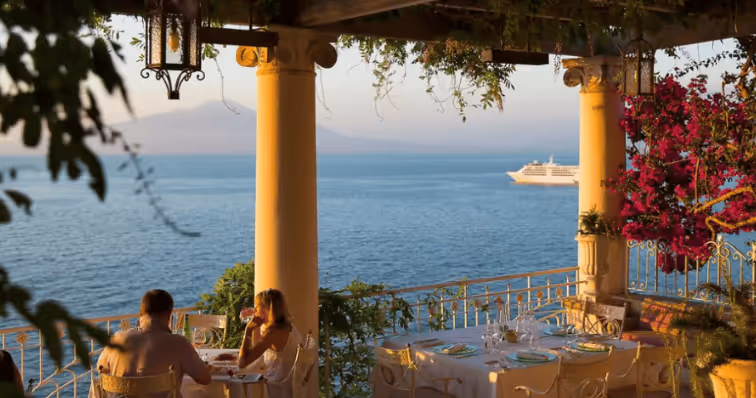 Couple dining at a table on a seaside terrace with columns, overlooking a calm ocean with a cruise ship in the distance during sunset.