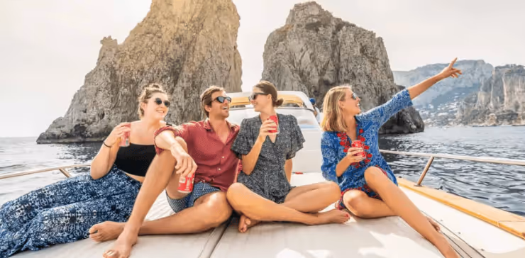 Four friends sitting on a boat deck, drinking soda and enjoying a sunny day near large rock formations in the sea.