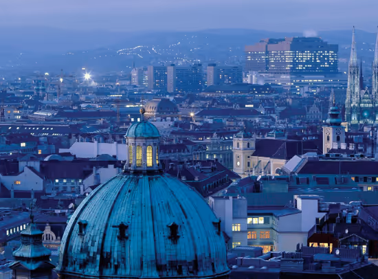 Cityscape at dusk with a large greenish-blue domed building in the foreground and various illuminated buildings and spires in the background.