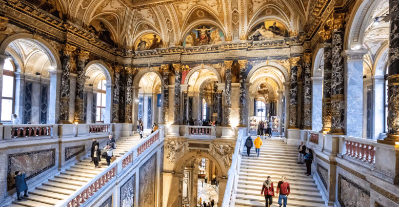 Grand interior of a historic building featuring ornate marble columns, decorated vaulted ceilings, and two wide staircases with people walking up and down.
