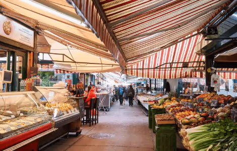 Busy covered market alley with fresh produce stands and seafood counters under striped awnings.