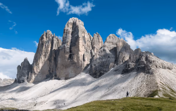 Jagged rocky mountain peaks under a blue sky with scattered clouds, with two tiny hikers standing on green grass in the foreground.