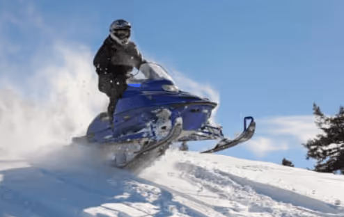 Person wearing a helmet riding a blue snowmobile over a snowy hill with trees and a clear sky in the background.