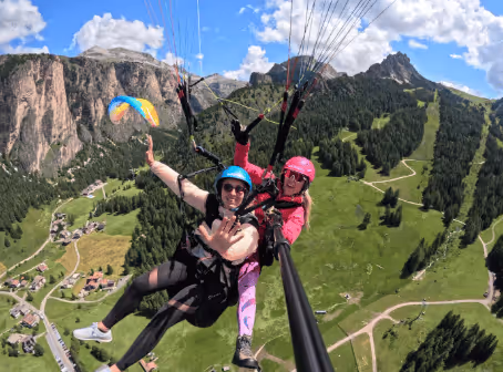 Two people tandem paragliding over a green mountainous landscape with rocky cliffs and a colorful parachute in the background.