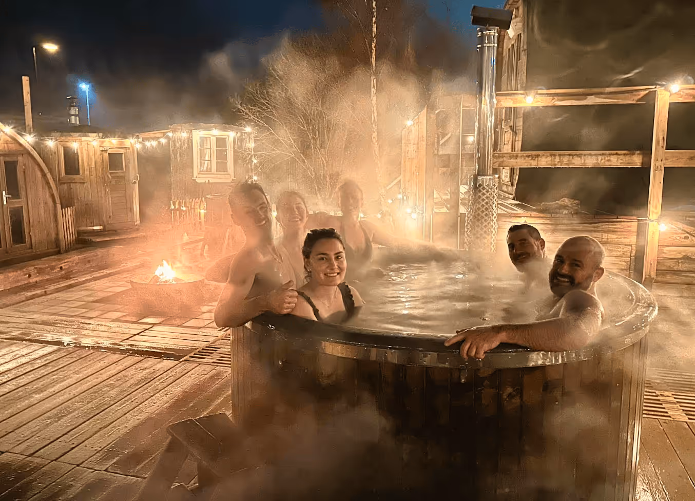 Six people smiling and relaxing in a steaming wooden hot tub at night with warm outdoor lighting.