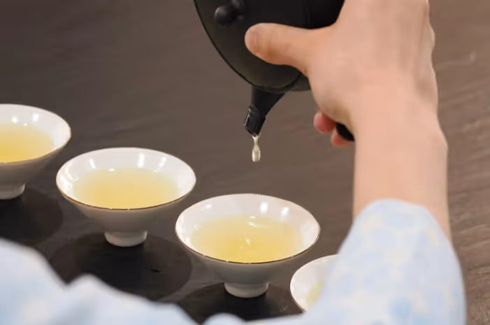 Hand pouring tea from a black teapot into small white ceramic cups with light yellow tea on a wooden surface.