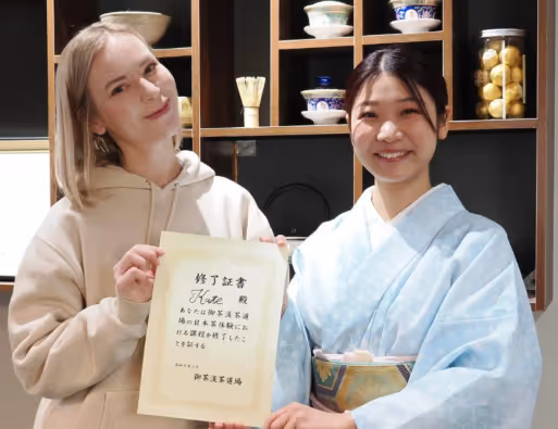 Two women smiling indoors, one in a beige hoodie and the other in a light blue traditional kimono holding a certificate with Japanese text.