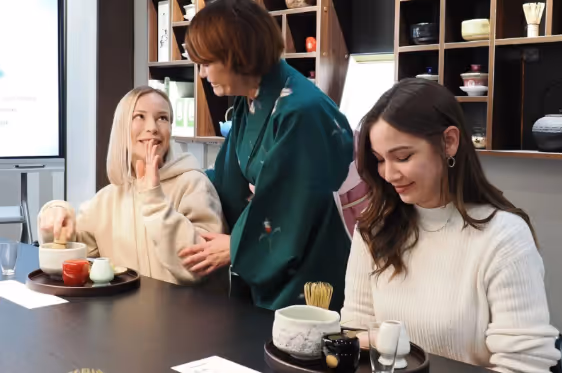 Three women sharing a moment while preparing and enjoying tea at a wooden table with tea utensils.