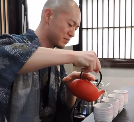 A man wearing a traditional Japanese robe pouring tea from a red teapot into white cups.