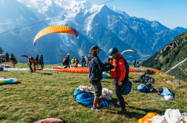 People preparing for paragliding on a grassy mountain slope with snow-capped peaks in the background.