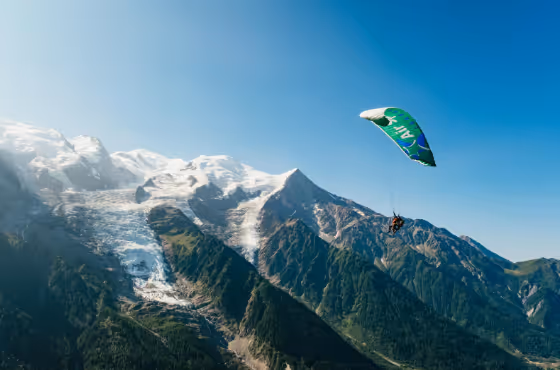 Paraglider flying over green mountains with snow-capped peaks under a clear blue sky.