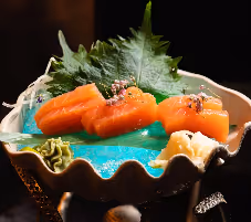 Three pieces of salmon nigiri sushi garnished with sesame seeds, served on a blue plate with green shiso leaf and wasabi.