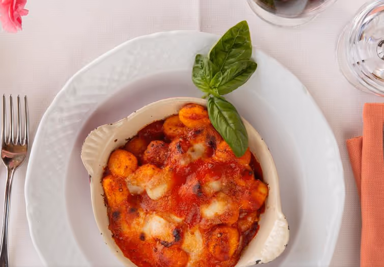 Baked gnocchi with tomato sauce and melted cheese garnished with a fresh basil leaf in a white dish on a white plate.