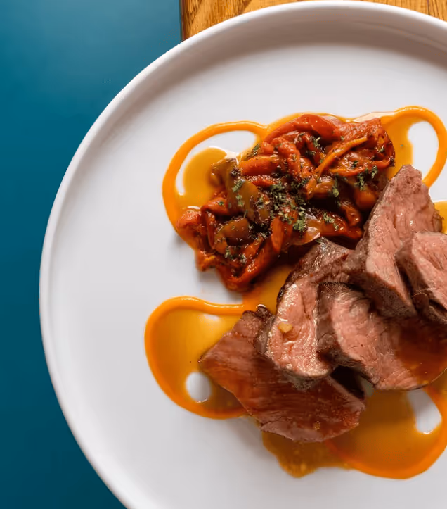 Plate with slices of medium-rare steak and roasted red peppers with sauce on a white plate against blue and wooden background.