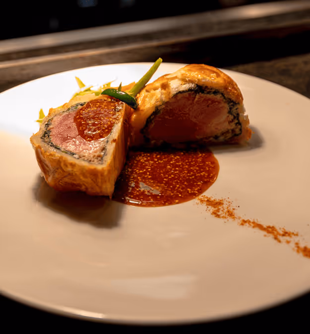 Plate with two halves of a beef Wellington topped with sauce and garnished with a green bean and vegetables.