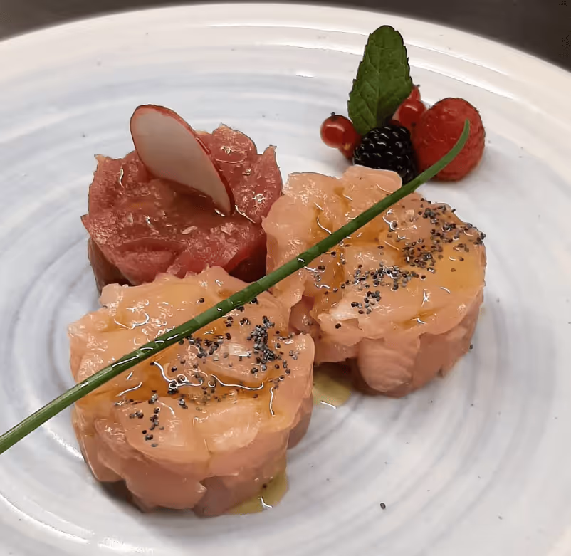Gourmet dish with three round tartare servings topped with sauce and poppy seeds, garnished with a chive stalk, mint leaf, and mixed berries on a white plate.