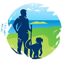 Silhouette of a person holding a walking stick standing next to a dog, with a scenic background of blue sky, green grass, and distant hills.